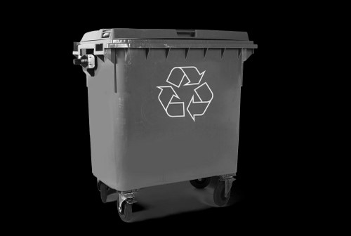 A medium-sized grey plastic waste bin with a hinged lid, prominently displaying a white recycling symbol on the front, situated on a flat, dark surface. The bin has four small caster wheels at the base, allowing for easy manoeuvring. The surrounding environment appears plain and unadorned, with no additional objects or background context visible, emphasizing the bin’s sturdy, textured surface. The lighting highlights the contours of the lid and the recycling logo, reflecting typical waste management equipment used by commercial waste removal services in Tottenham. The bin is positioned upright, with the lid closed, ready for collection or disposal, and its neutral colour and design suggest suitability for handling a variety of rubbish types in a professional waste management setting, such as those operated by Commercial Waste Tottenham in the London postal area.
