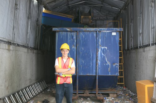 A worker wearing a yellow hard hat, high-visibility safety vest with reflective stripes, a white shirt, and dark trousers is standing with arms crossed in front of a large, blue industrial waste container situated inside a metal-framed waste storage facility. The container has a weathered surface with visible scratches and dents, and is positioned on a concrete floor scattered with small debris and rubbish. To the right of the container, there is a yellow safety barrier or bin, and behind the container, a yellow ladder is leaning against the structure's wall. The interior walls and ceiling of the facility are made of metal, with visible supporting beams and panels, and the lighting appears natural, possibly from open doors or windows. The scene is set in an industrial or commercial waste management environment, consistent with rubbish removal services in Tottenham, with the focus on waste containment and professional staff readiness.