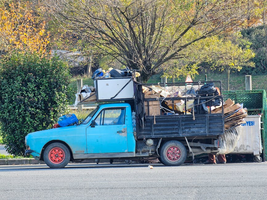 A small, old blue flatbed truck parked on the side of a paved street, heavily loaded with various waste materials including large cardboard boxes, plastic bags, and miscellaneous debris. The truck's p