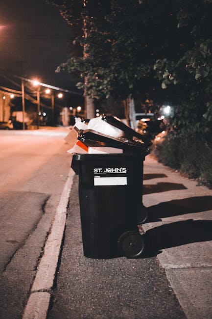 A small, old blue flatbed truck parked on the side of a paved street, heavily loaded with various waste materials including large cardboard boxes, plastic bags, and miscellaneous debris. The truck's p