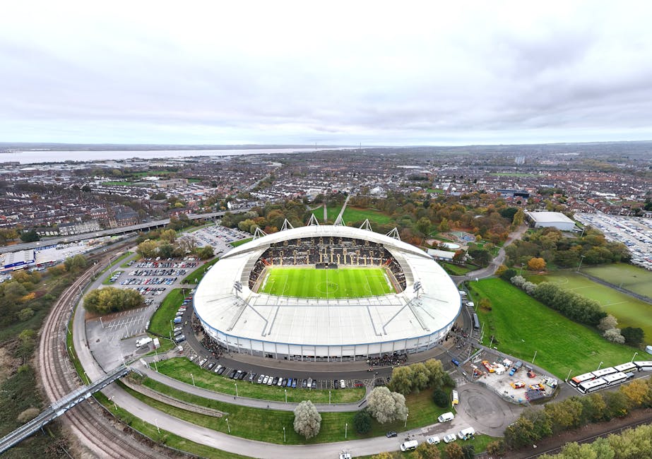 An aerial view of the White Hart Lane stadium, with its large, oval-shaped white roof and exterior walls made of metallic panels. The main entrance features three prominent gate structures beneath the